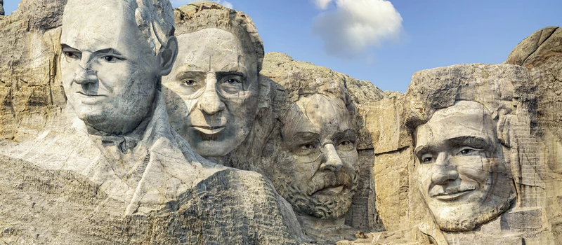 Vier Männergesichter als Felsrelief im Stil von Mount Rushmore bei blauem Himmel.