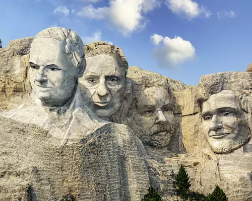 Vier Männergesichter als Felsrelief im Stil von Mount Rushmore bei blauem Himmel.